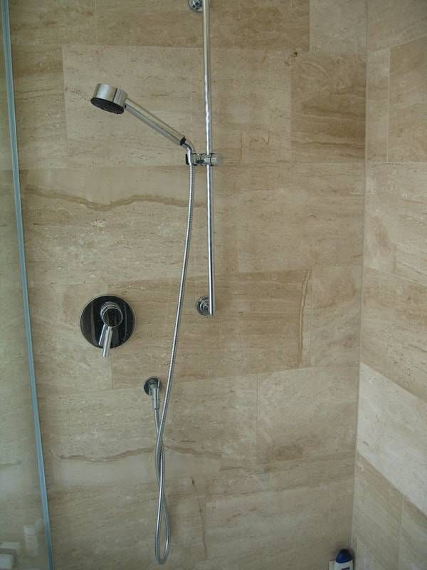 Eine Handbrause, die an einer verschiebbaren Metallstange an einer beigefarbenen, rechteckigen Fliesenwand in einer Duschkabine befestigt ist.