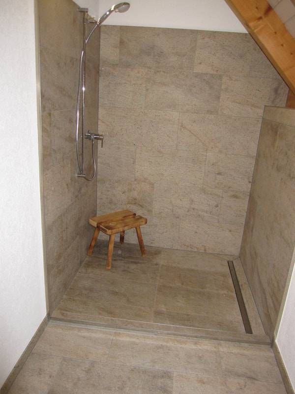 Eine ebenerdige Dusche mit beigefarbenen Steinfliesen, einem Holzhocker und einem Edelstahlablauf in einem Badezimmer mit Dachschrägen.