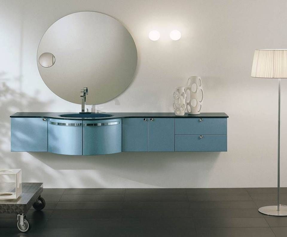 Ein moderner, blauer, schwebender Waschtisch mit rundem Spiegel und Stehlampe vor einer weißen Wand.