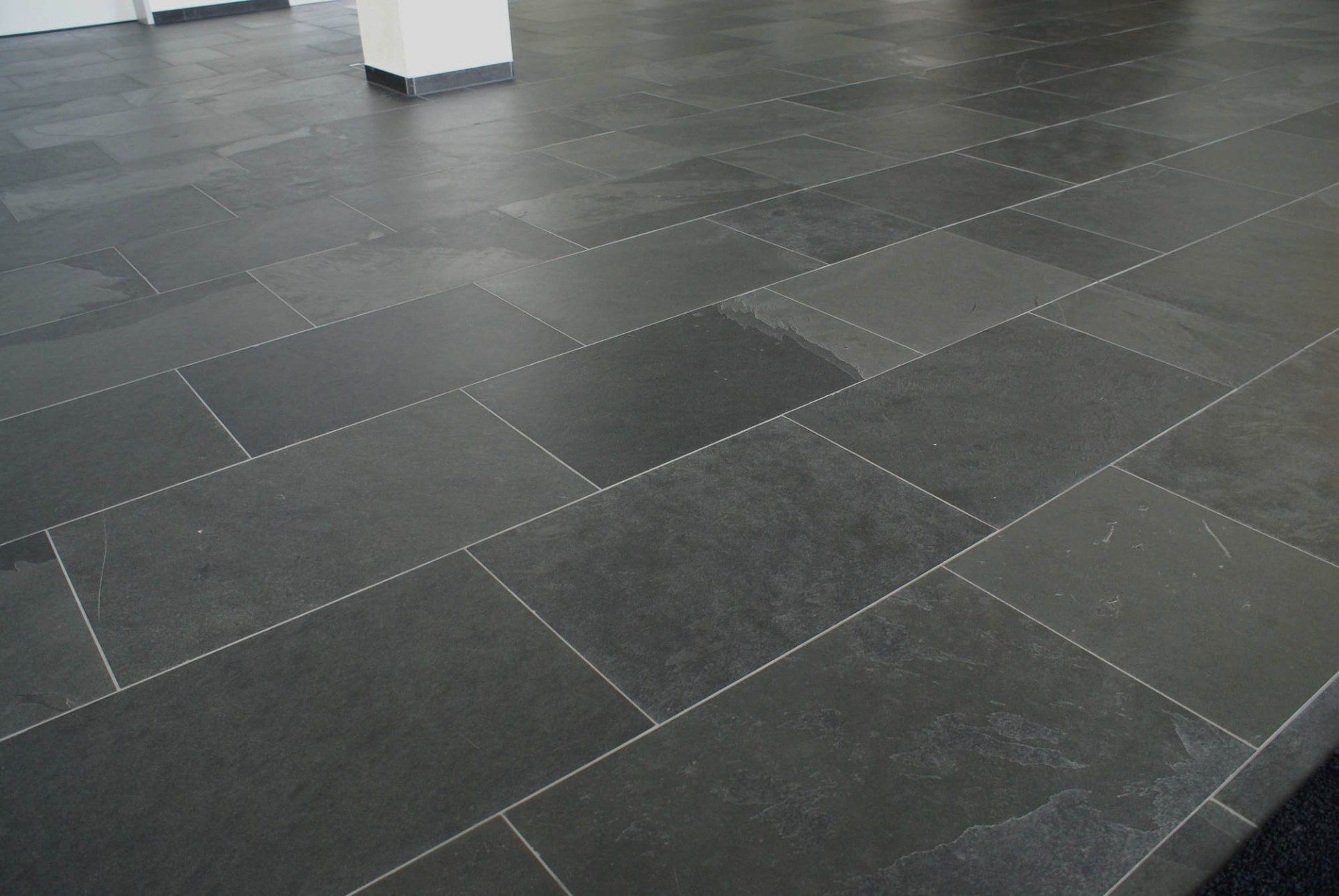 Ein Boden, der mit rechteckigen, dunkelgrauen Steinfliesen belegt ist, die ein sichtbares Raster aus hellen Fugenlinien aufweisen.