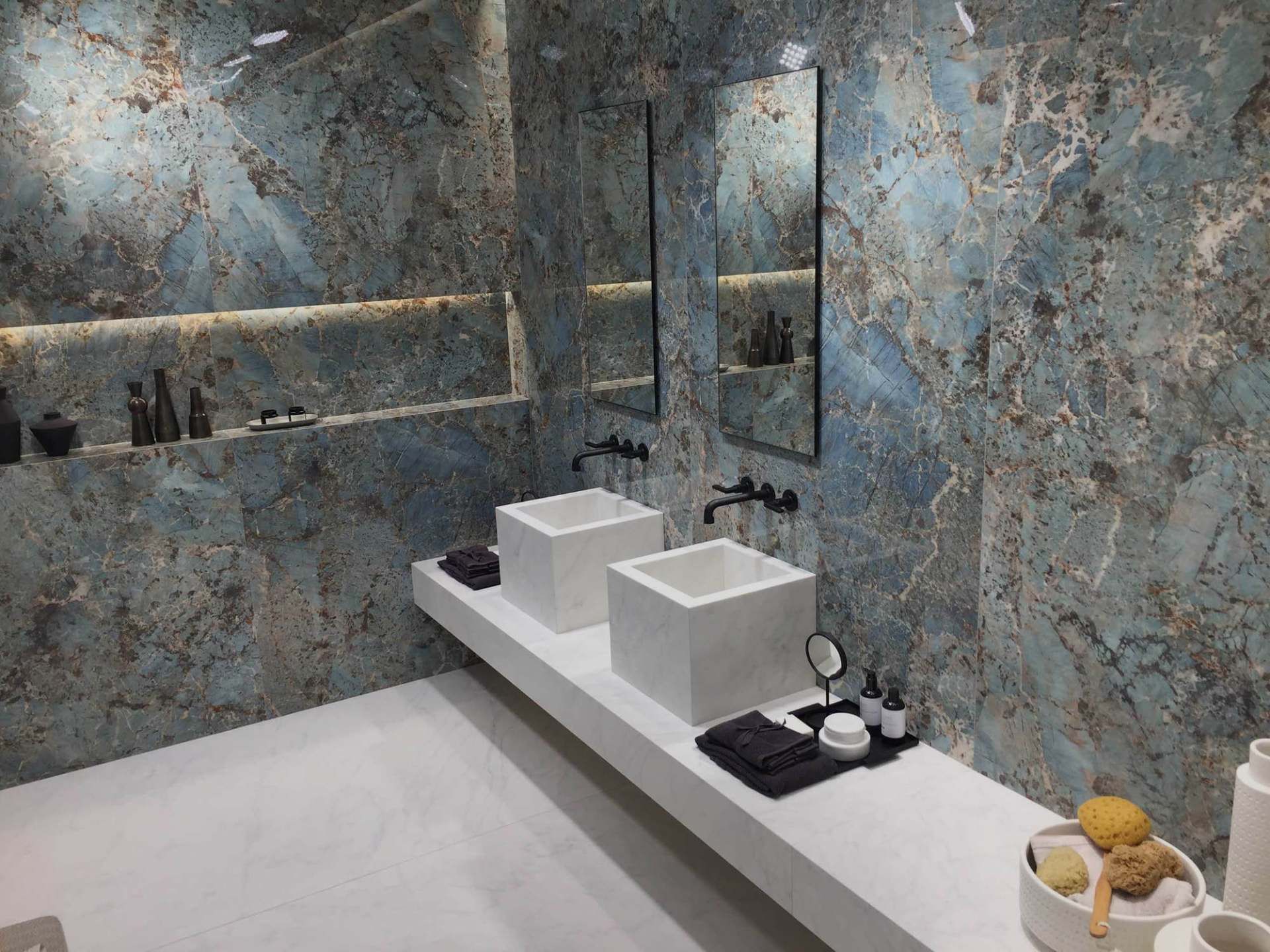 Ein modernes Badezimmer mit blaugrauen Marmorwänden, zwei weissen quadratischen Waschbecken auf einem weissen, schwebenden Waschtisch und zwei Spiegeln.