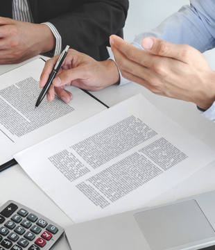 Deux personnes examinent des documents à un bureau : l'une pointe du doigt, l'autre écrit au stylo. Une calculatrice.