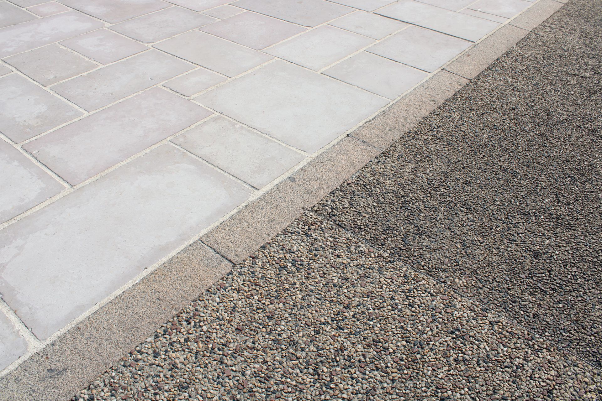 Une allée pavée de pierre claire, séparée d'une surface de gravier foncé texturée par une bordure en béton lisse.