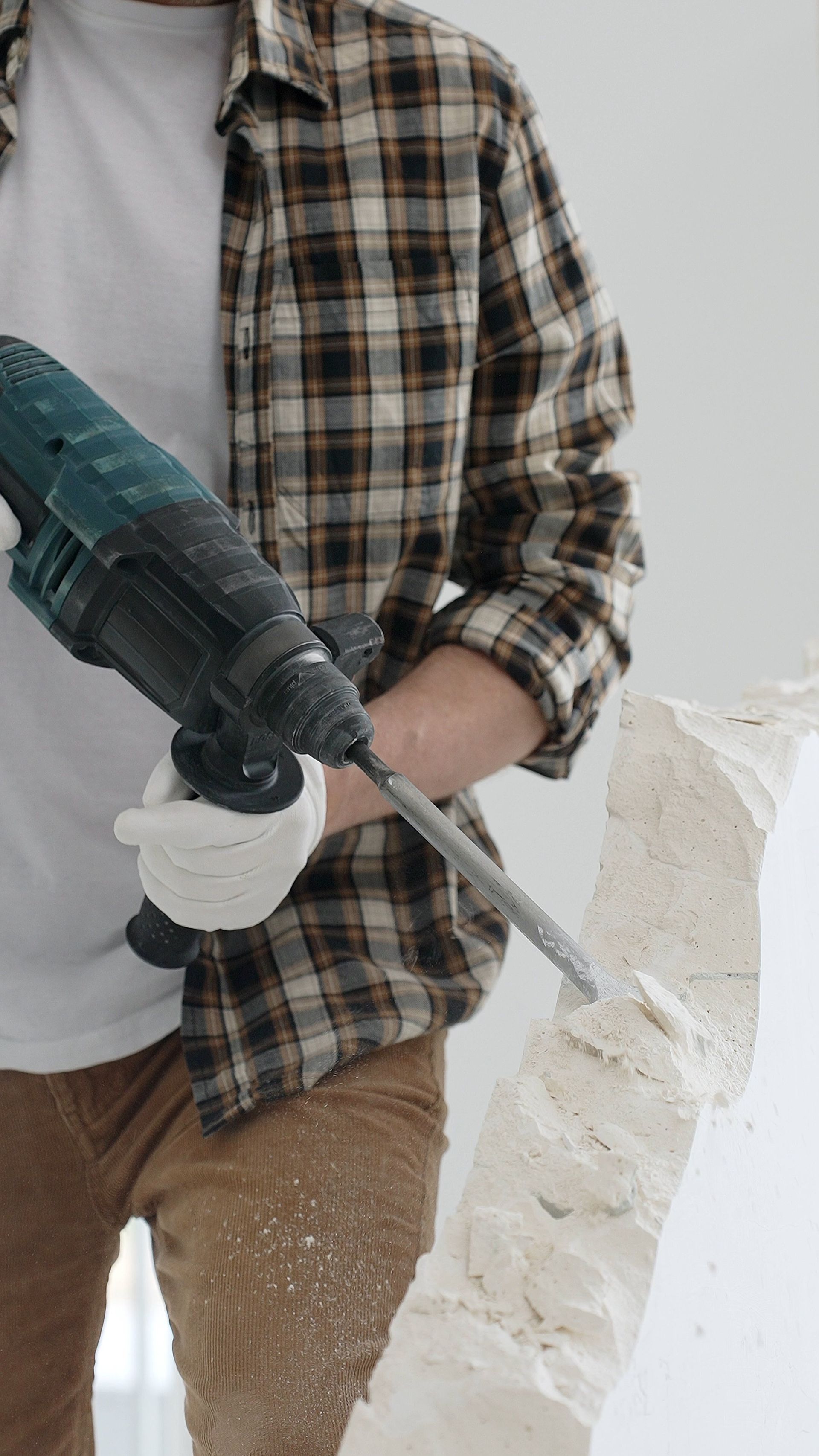 Une personne portant une chemise à carreaux et des gants utilise un marteau perforateur pour casser un gros bloc de béton.