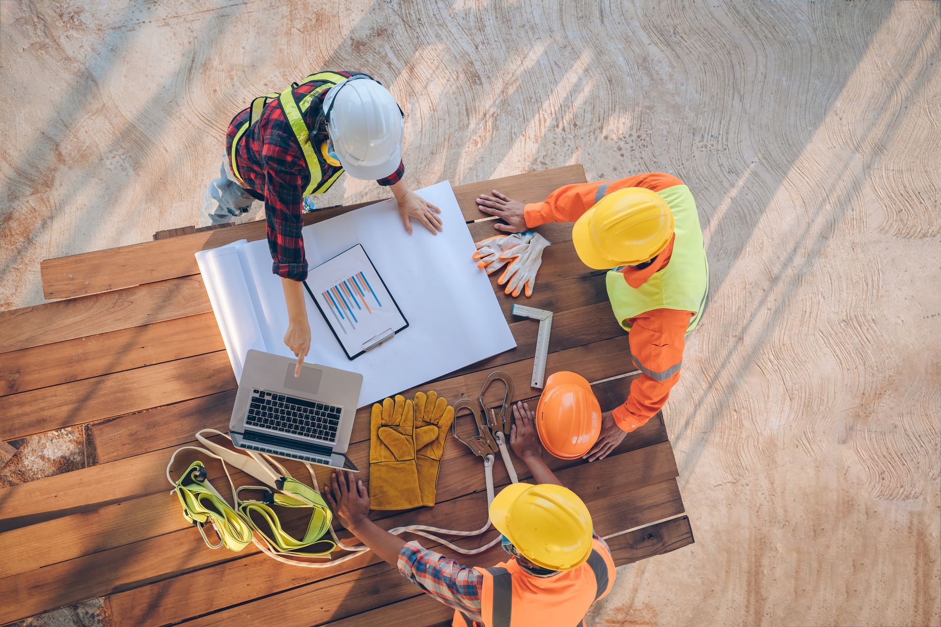 Sur un chantier, trois ouvriers portant des gilets haute visibilité et des casques de sécurité examinent des plans et un ordinateur portable posés sur une surface en bois.