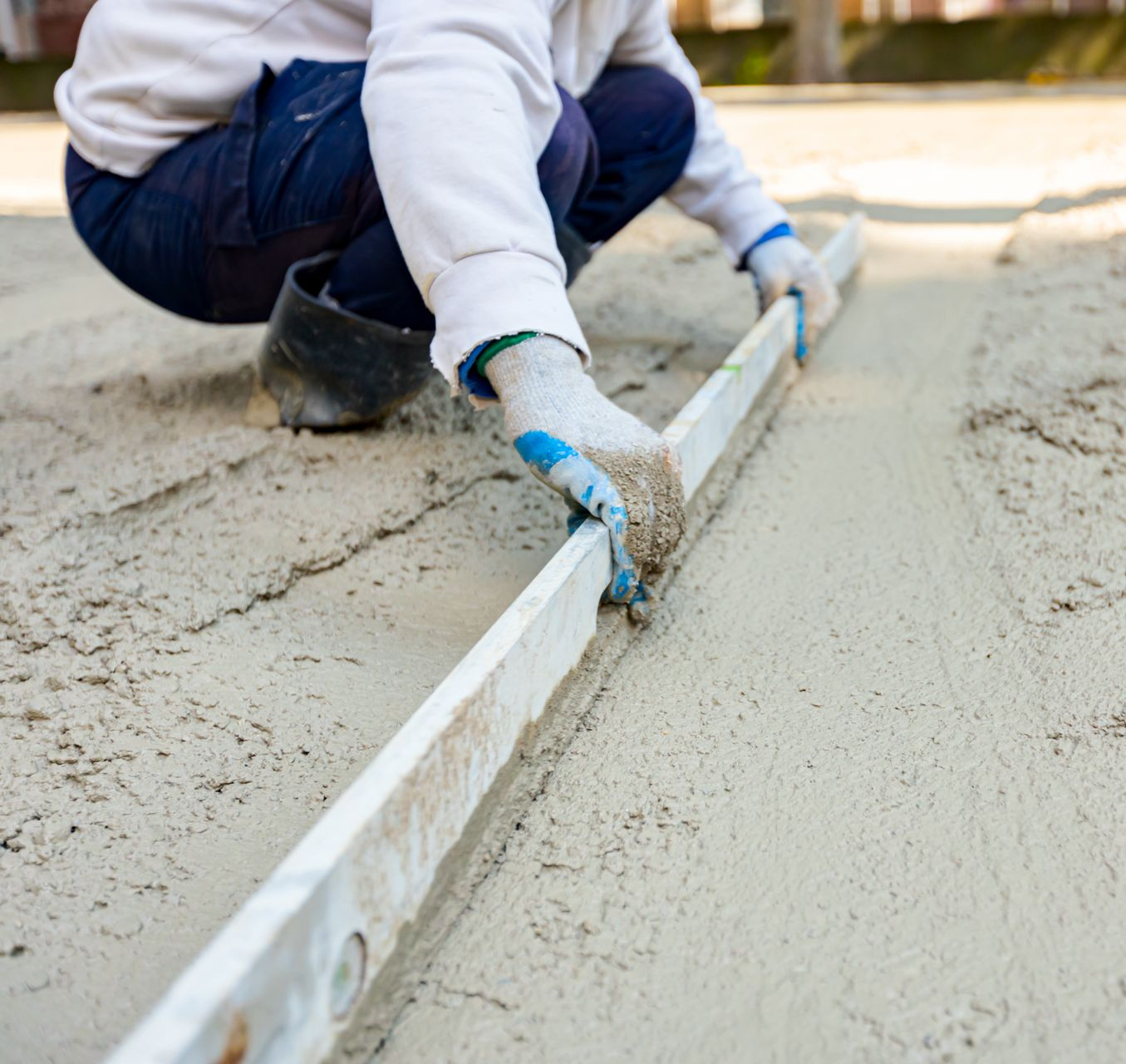 Une personne portant des vêtements et des gants de travail utilise un outil de nivellement en métal pour lisser le béton fraîchement coulé sur le sol.