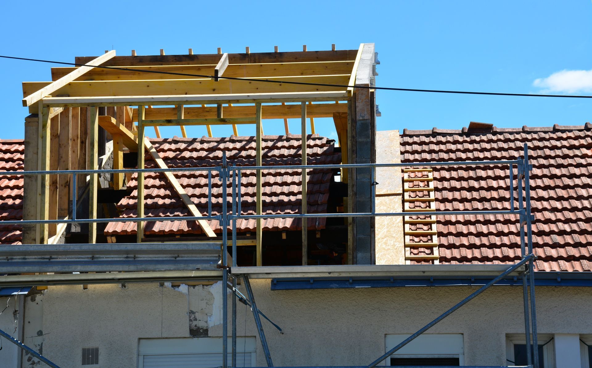 Une charpente de lucarne en bois en construction sur un toit de tuiles rouges, se détachant sur un ciel bleu dégagé.
