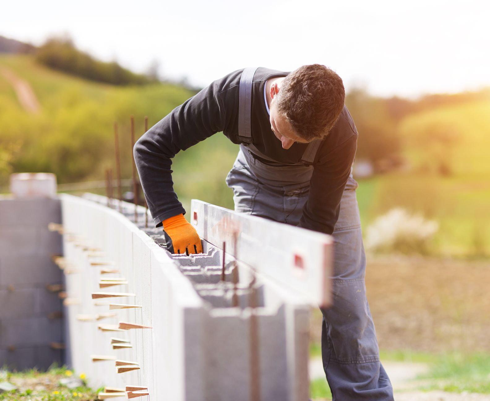 Un ouvrier en combinaison grise et gants orange utilise un niveau à bulle pour vérifier l'alignement d'un mur en blocs de béton.