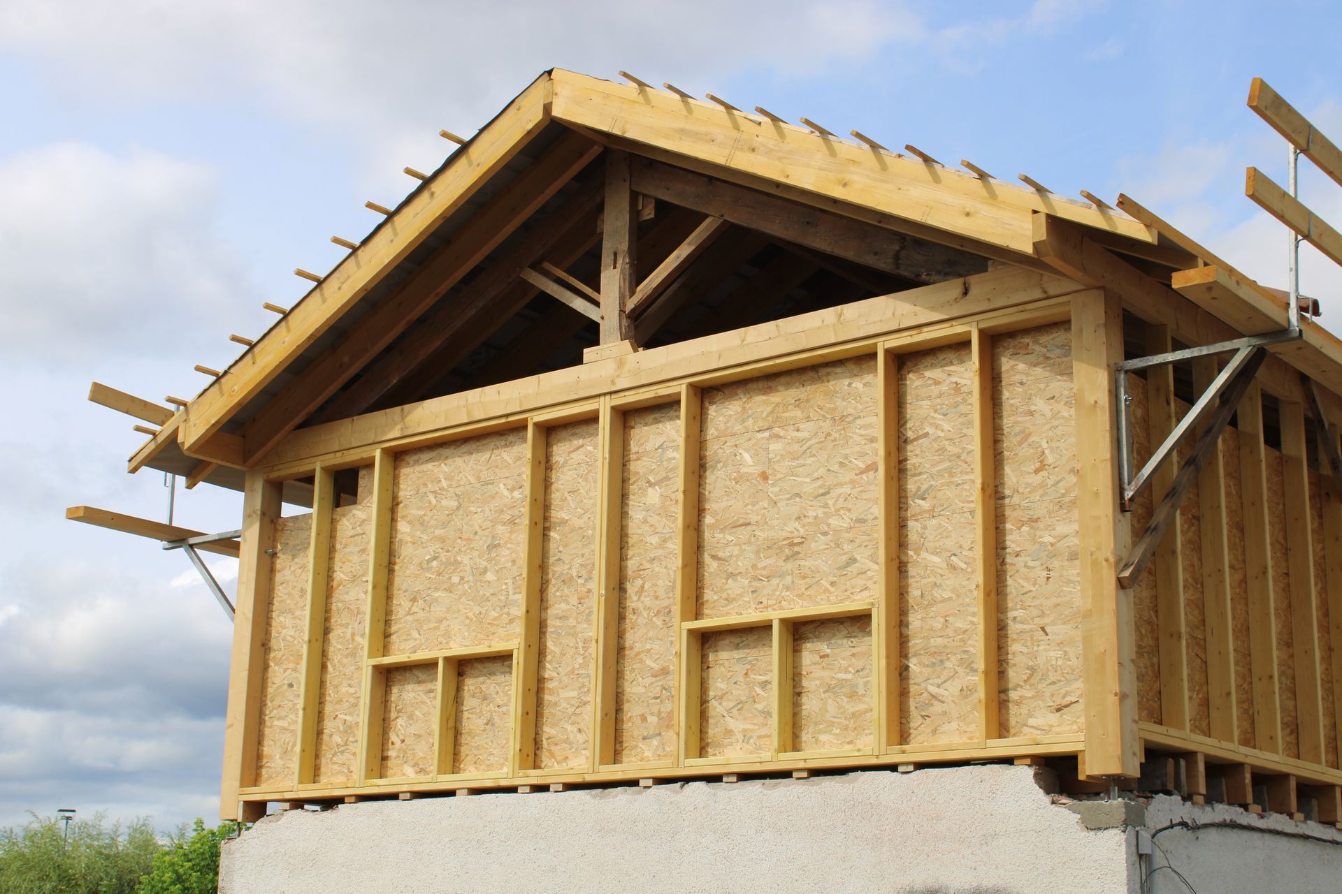 Une maison en construction, avec une charpente en bois apparente et des panneaux OSB sous un toit à pignon, se détachant sur un ciel nuageux.