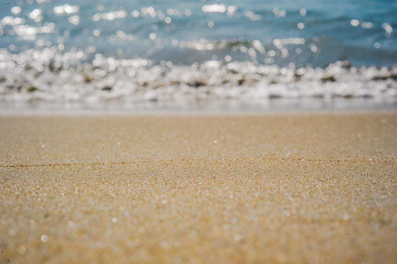 a low angle view of sparkling sand with soft blurred water and sunlight in the background, symbolising the quiet beauty of moving through life as an autistic woman in your own time and space
