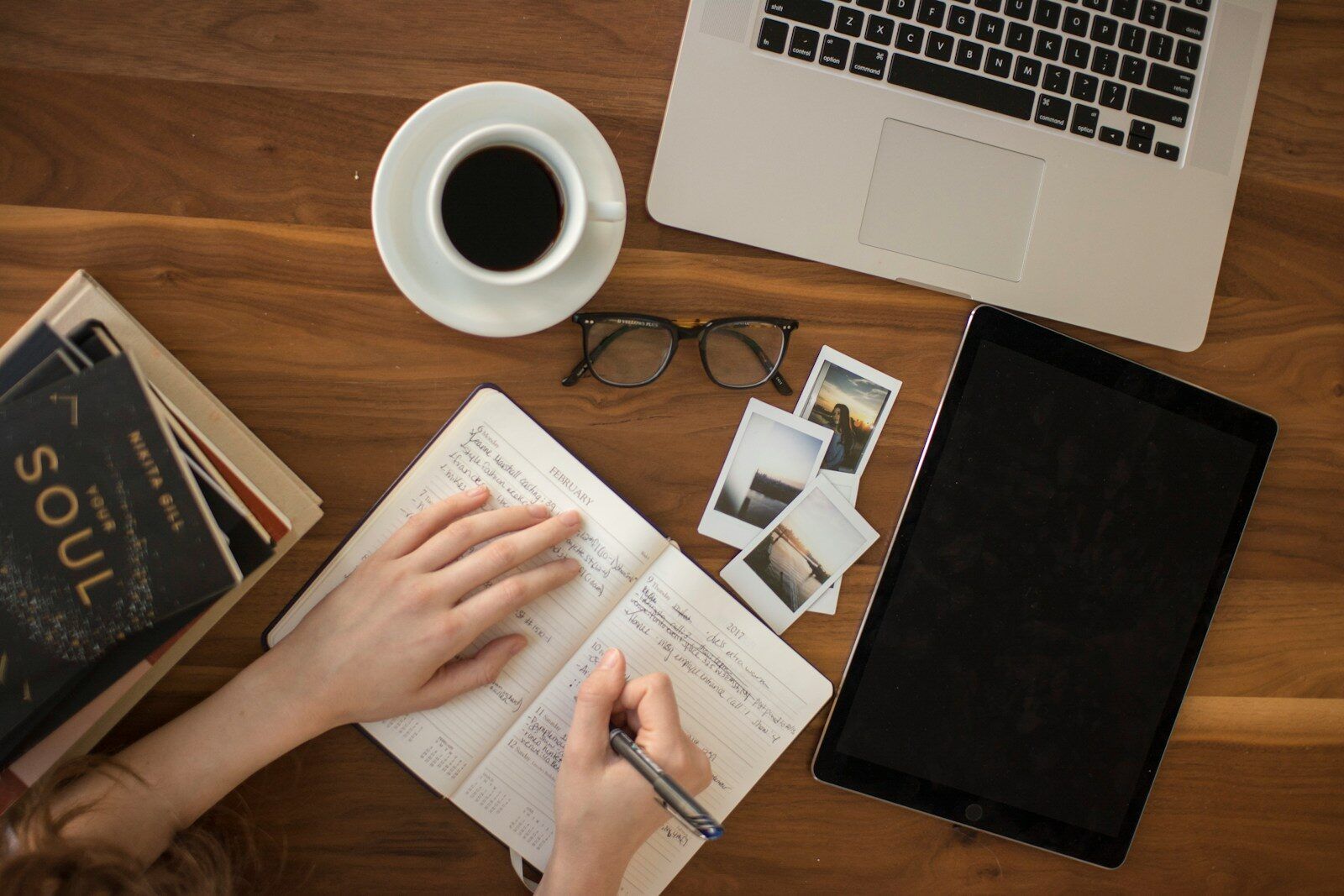 a bird's eye view of a desk with an open laptop, glasses, books, polaroid photos and a hand writing in a notebook, representing the process of reflection and understanding after a late neurodivergent diagnosis