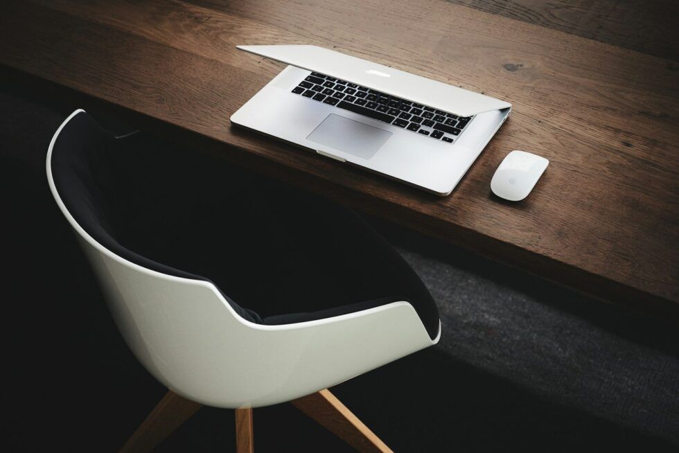 a dimly lit wooden desk with an empty chair and a half-closed laptop, symbolising the cost of exclusion and the exhaustion of neurodivergent people in environments not built for them