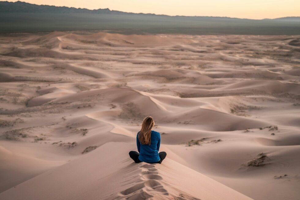 a woman sitting cross-legged on sand dunes looking into the distance, symbolising reflection and post-diagnosis grief in neurodivergent adults