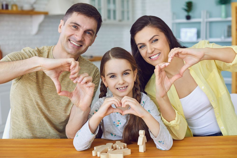 Una familia sonríe mientras forma corazones con las manos sobre un juego de juguetes de madera colocado sobre una mesa en una cocina luminosa.