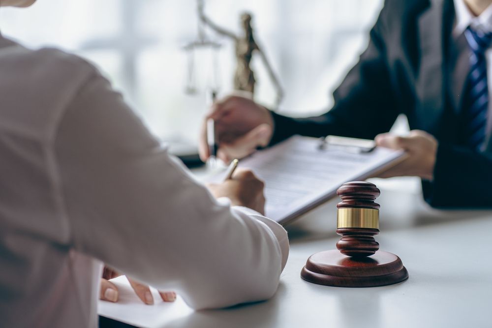 Un abogado y su cliente revisan un documento en un escritorio, con el mazo de un juez y una estatua de la Dama de la Justicia al fondo.