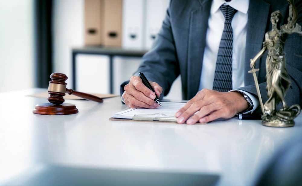 Un profesional vestido de traje escribe en un bloc de notas en un escritorio con un mazo de juez de madera y una estatua de la Dama de la Justicia.