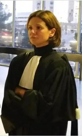 Femme en robe noire et col blanc, les bras croisés, debout à l'intérieur près d'une fenêtre.