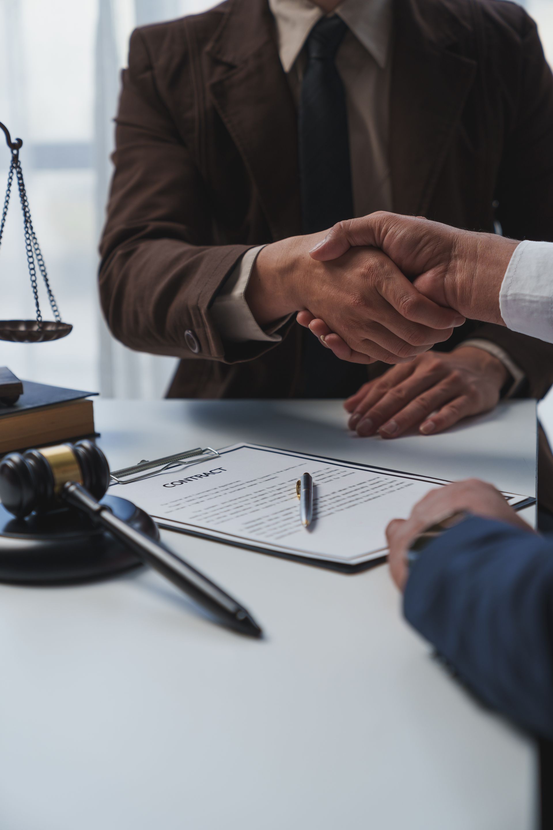 Deux personnes en costume se serrent la main au-dessus d'un document posé sur un bureau, avec un marteau et une balance de la justice visibles.
