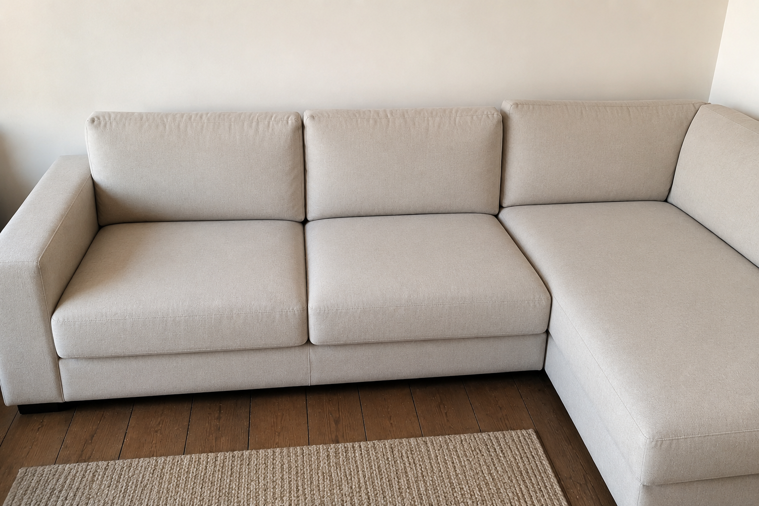 Canapé d'angle beige contre un mur clair, avec un tapis tissé sur un parquet.