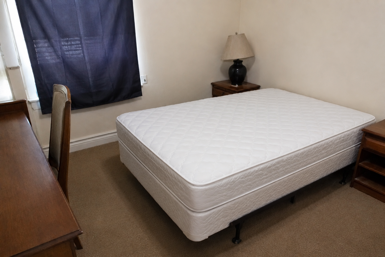 Petite chambre avec un lit blanc défait, un bureau en bois, une lampe et une fenêtre avec des rideaux sombres.