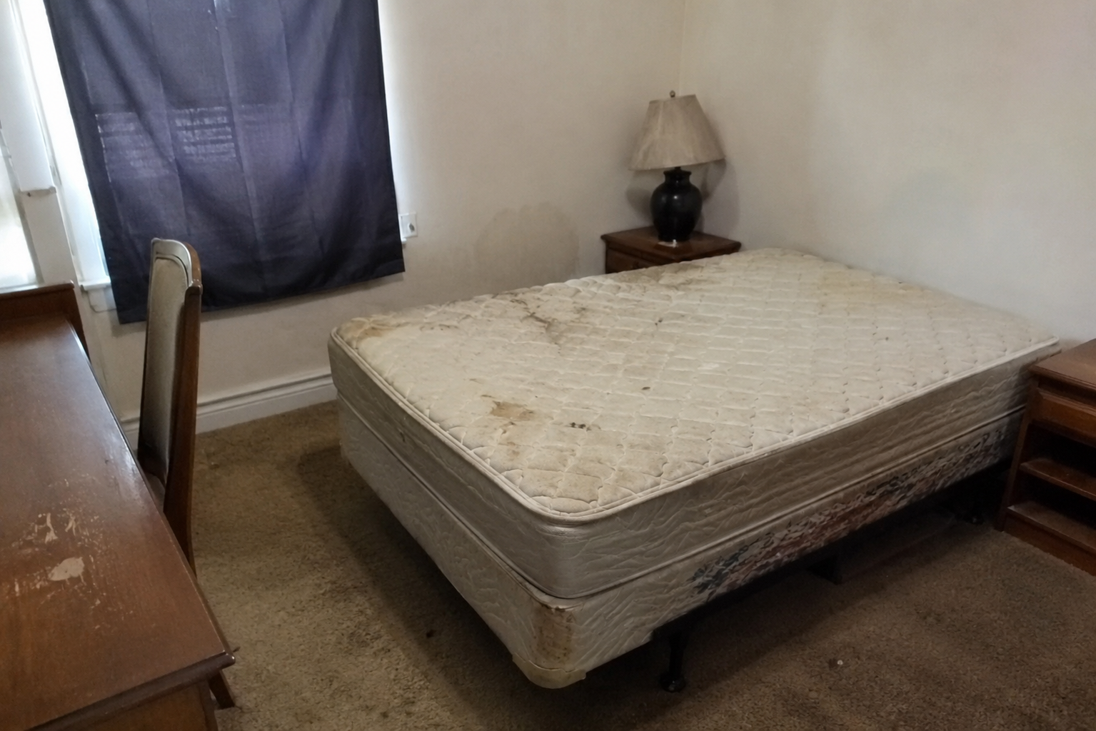 Petite chambre avec un lit défait, une lampe de chevet, une fenêtre et une commode sur des murs blancs.