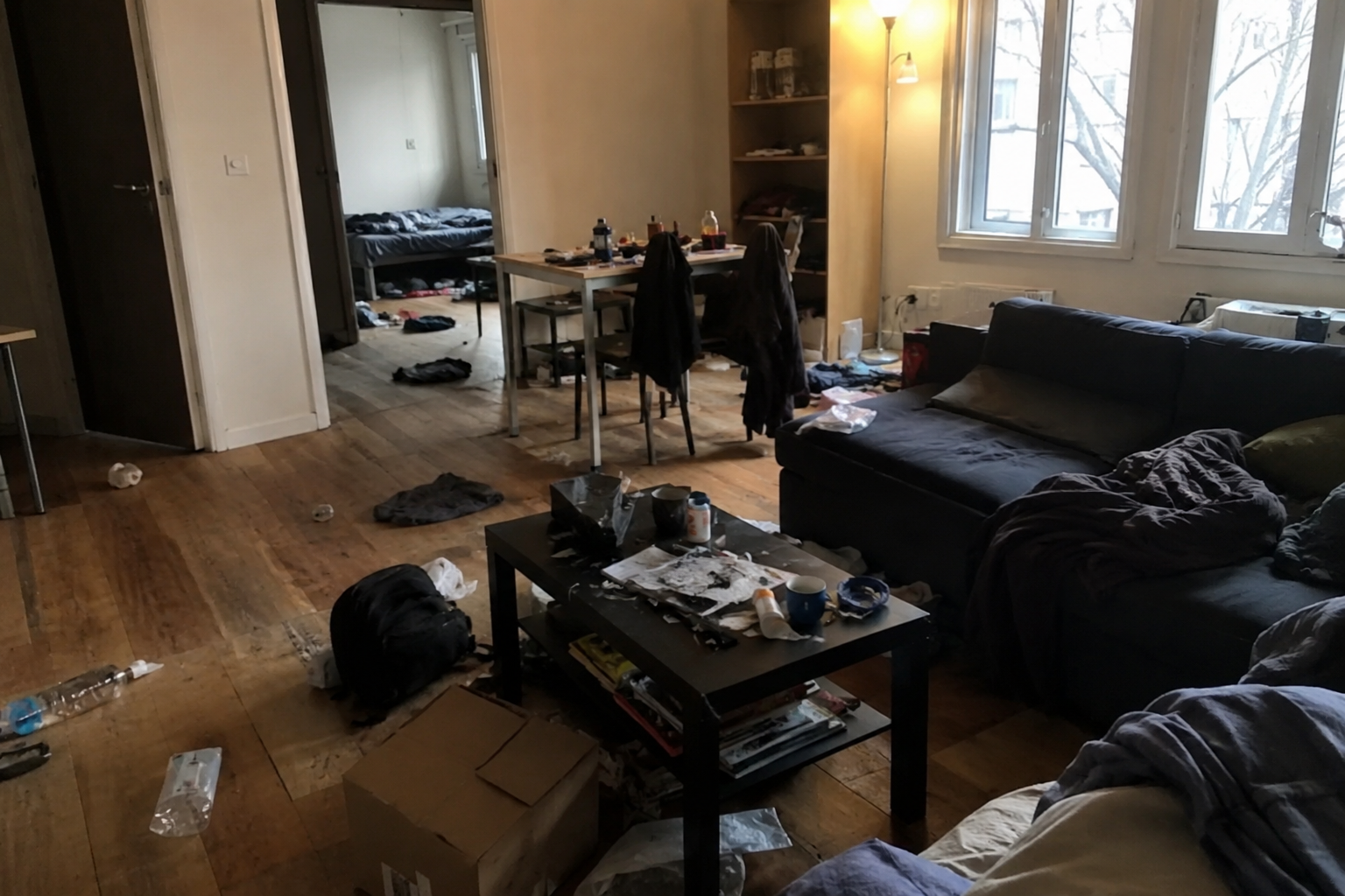 Salon encombré avec un canapé noir, une table basse, des cartons et la lumière du soleil entrant par une fenêtre.
