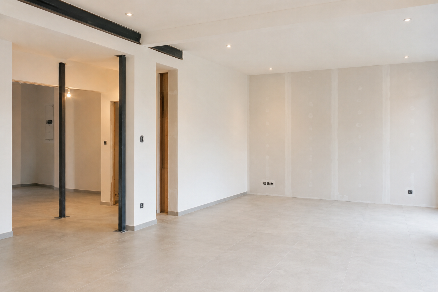Pièce moderne vide avec murs blancs, sol beige et colonnes et poutres noires apparentes.