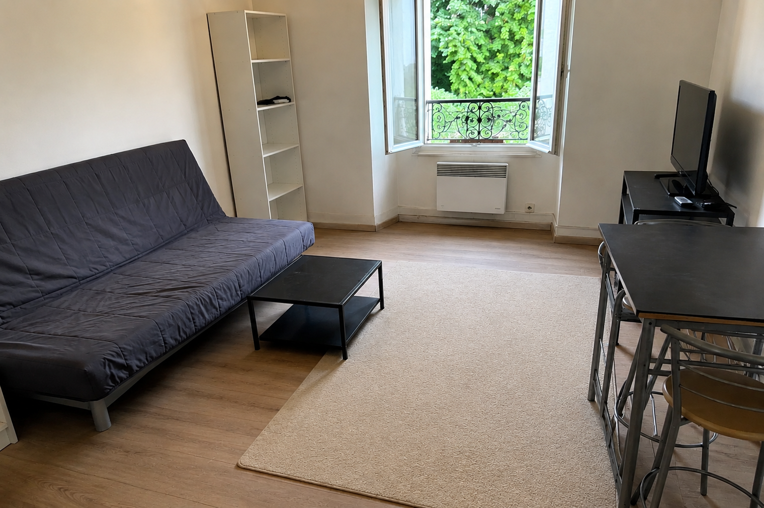 Petit salon avec un canapé foncé, une table basse, un bureau et une fenêtre ouverte donnant sur la verdure.