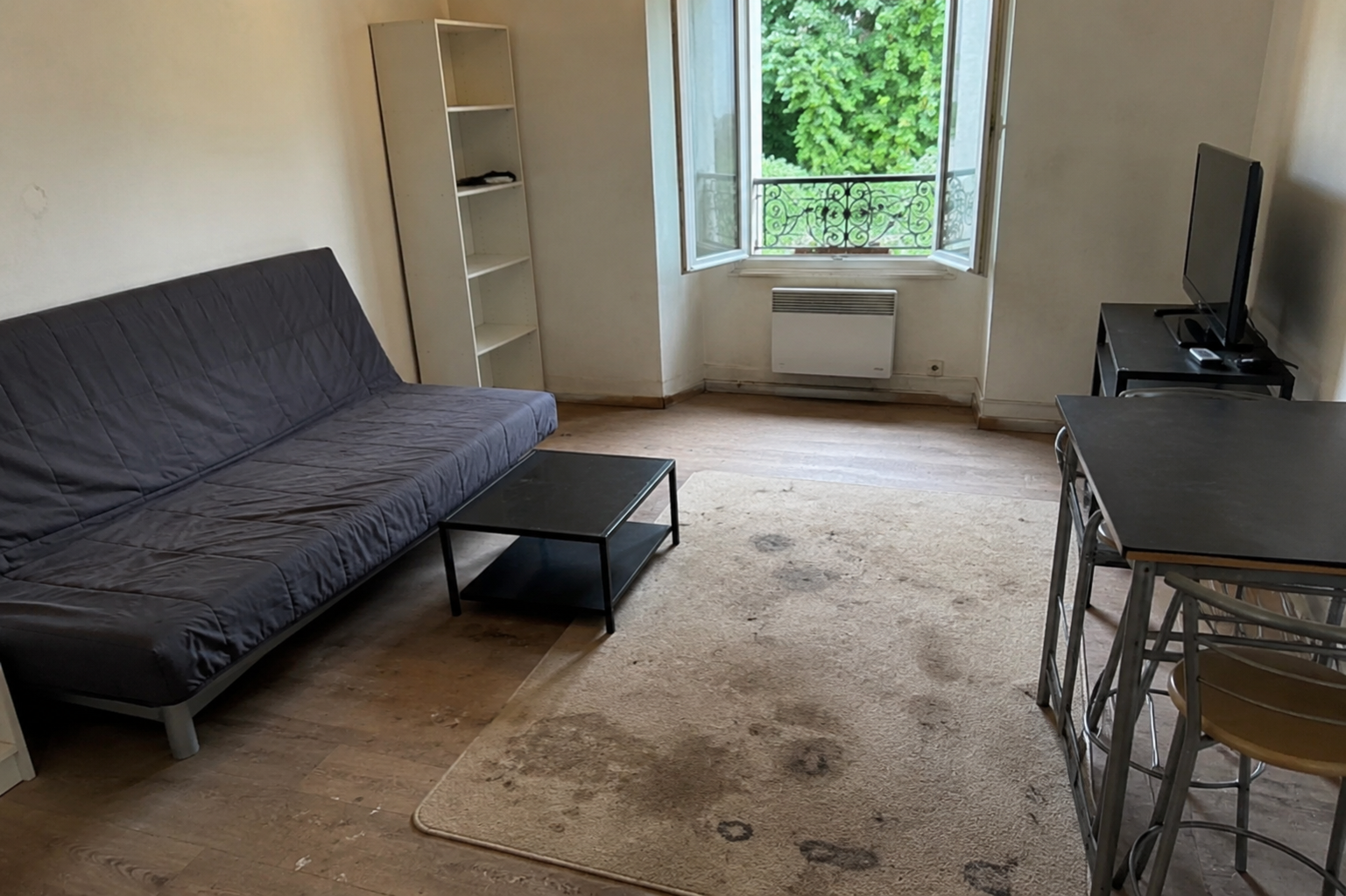 Salon épuré avec un canapé gris, une table basse, une télévision, une étagère et une fenêtre ouverte donnant sur la verdure.