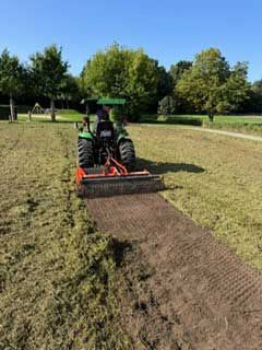 Ein Traktor mäht eine Wiese; im Hintergrund sind Bäume, ein Weg und ein Spielplatz unter blauem Himmel zu sehen.