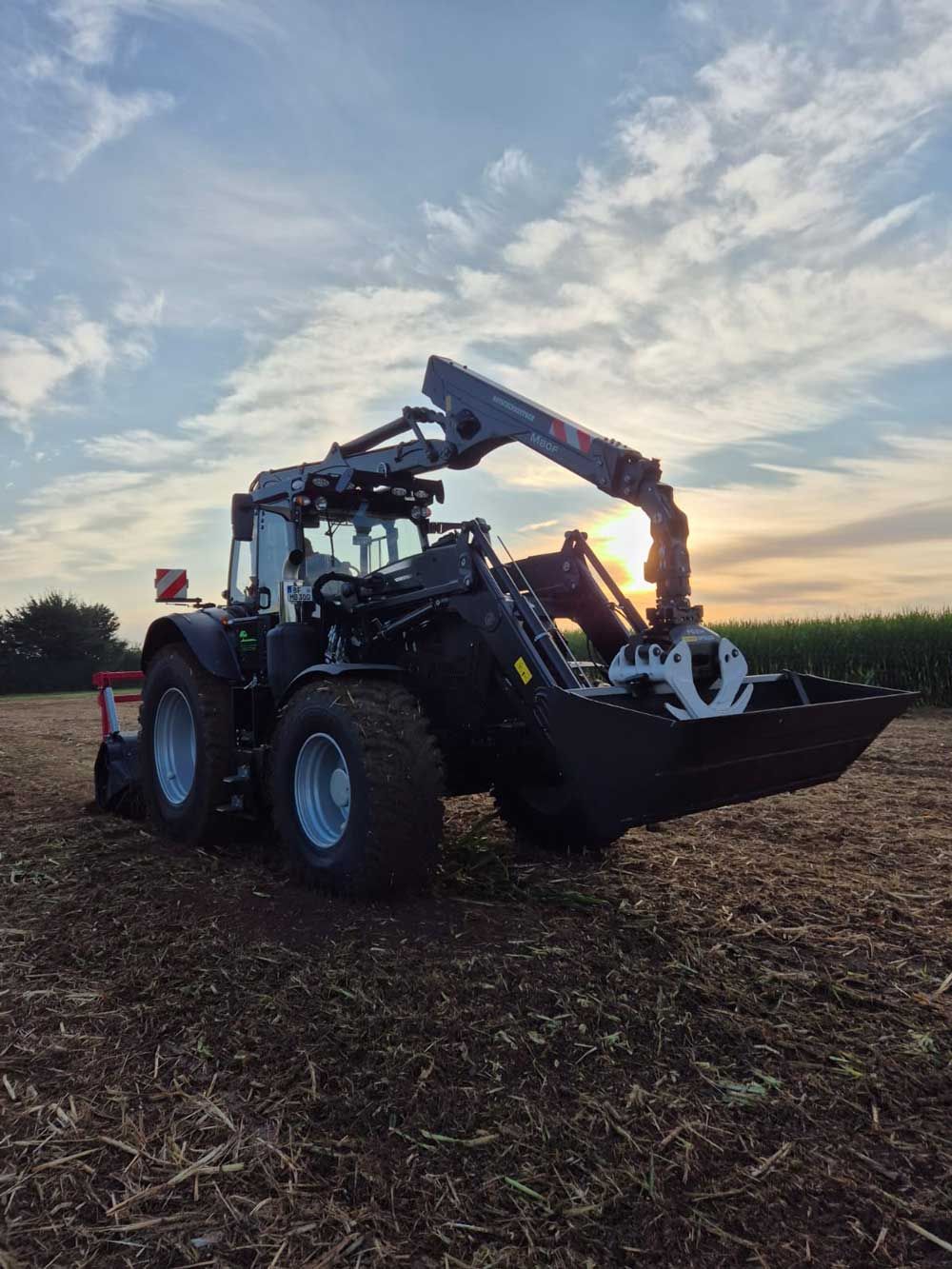 Traktor mit Frontlader und Greifer auf einem Feld bei Sonnenuntergang; als Silhouette vor einem bewölkten Himmel.