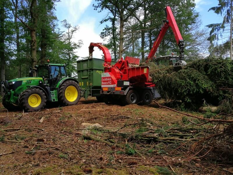 Ein grüner Traktor zieht einen roten Häcksler in einem Wald.