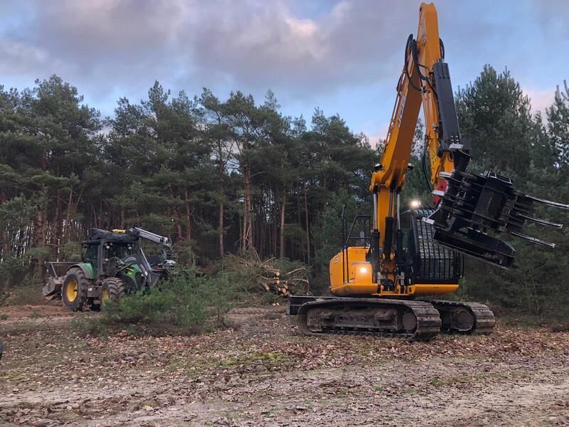 Ein großer gelber Bagger steht mitten im Wald neben einem Traktor.