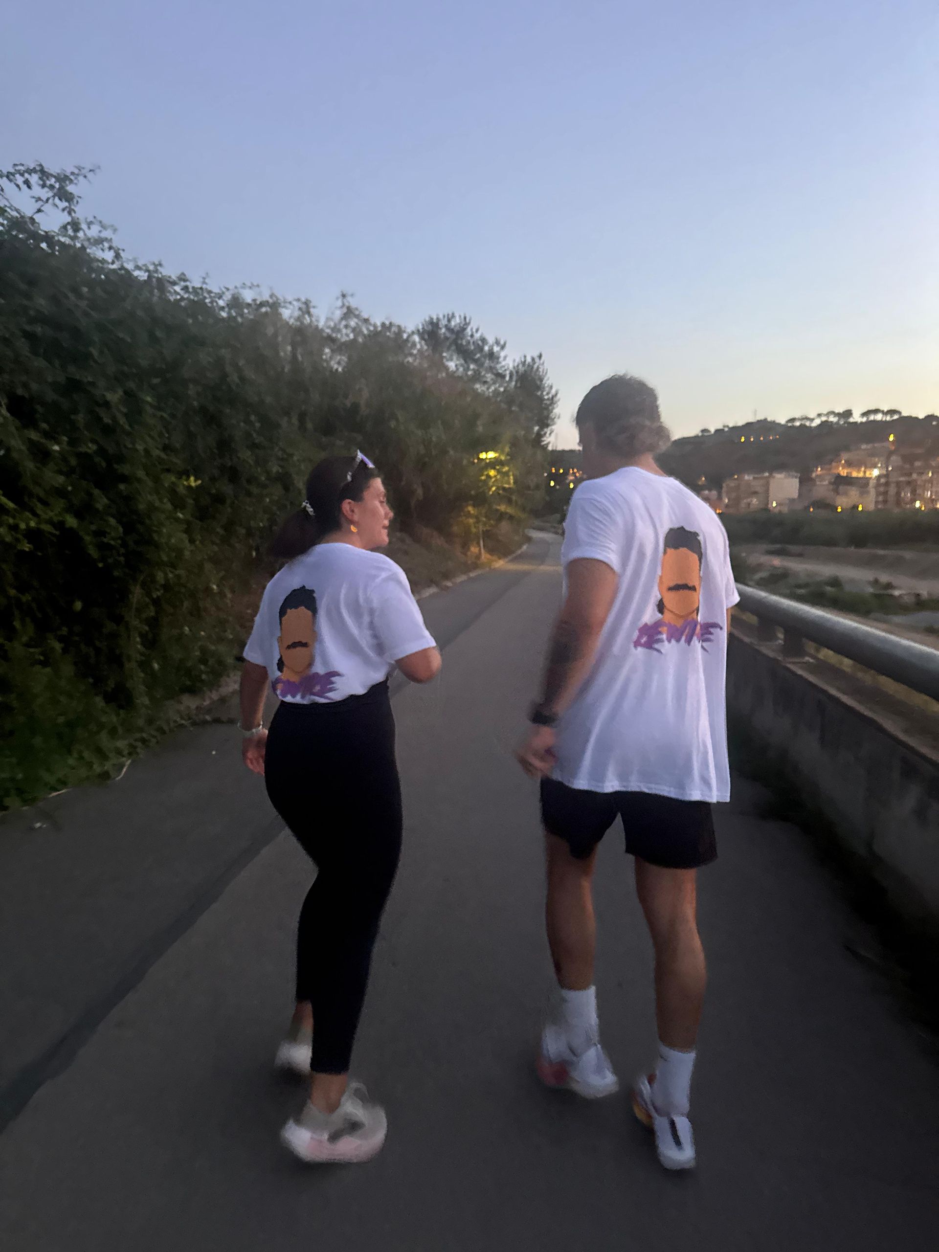 Dos personas caminan por un sendero, ambas con camisetas blancas con un gráfico en la espalda. Luz del atardecer.