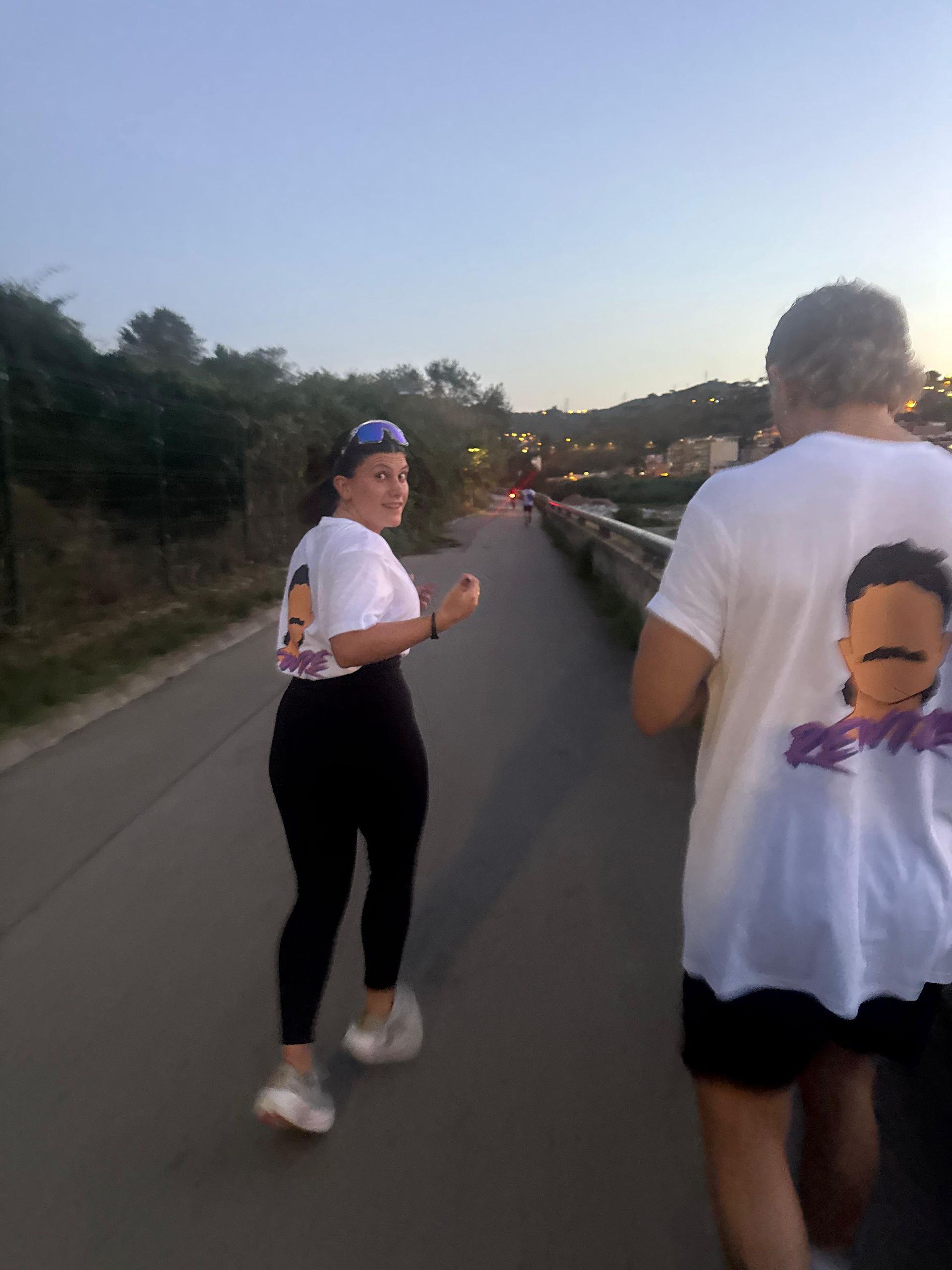 Dos personas vestidas con camisetas blancas y una cara de dibujos animados corren por un sendero al anochecer.