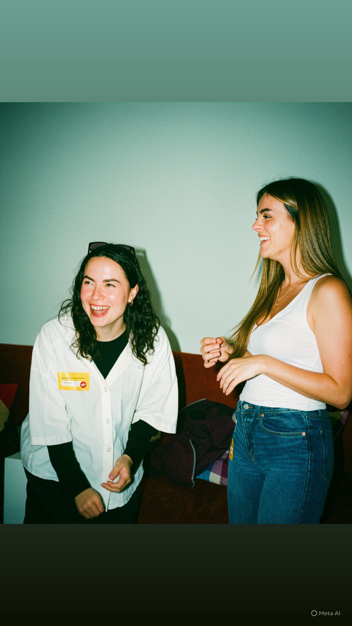 Dos mujeres riendo en una sala de estar, una sentada con una chaqueta blanca y la otra de pie con una camiseta blanca de tirantes y vaqueros.