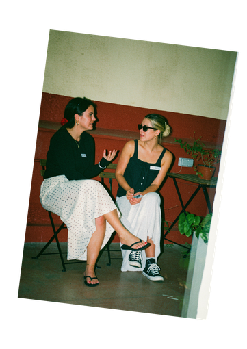 Dos personas sentadas en sillas conversando en una habitación sencilla; una de ellas lleva gafas de sol.