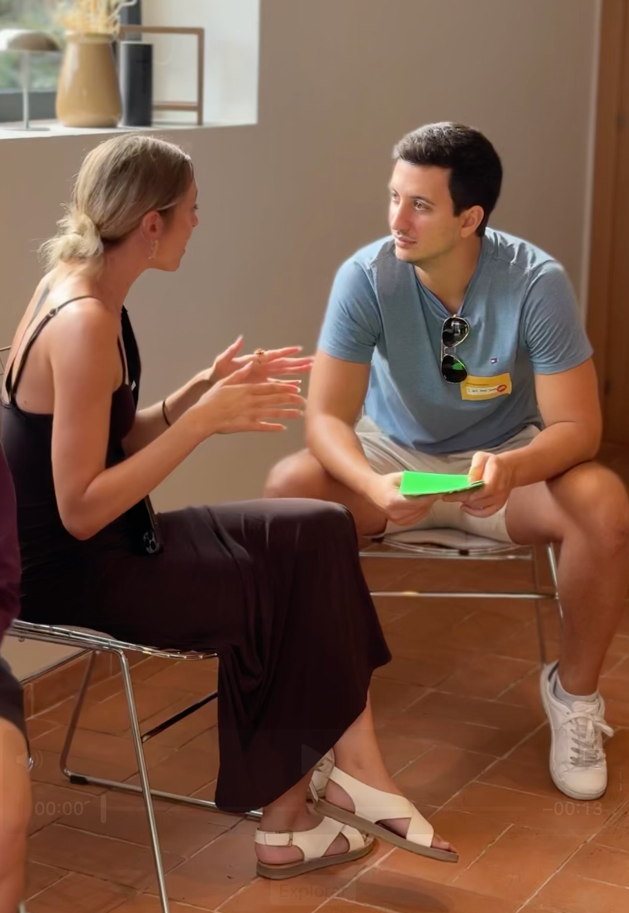 Dos personas conversando sentadas en sillas en una habitación luminosa; una de ellas sostiene una tarjeta de residencia permanente.