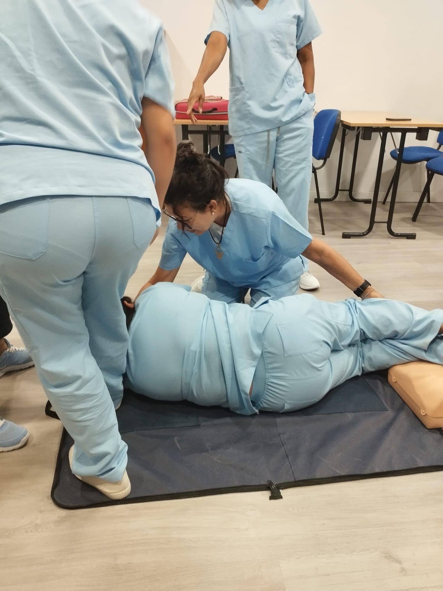Una cuidadora vestida de azul ayuda a dos ancianos a sentarse y conversar en una sala de estar luminosa.