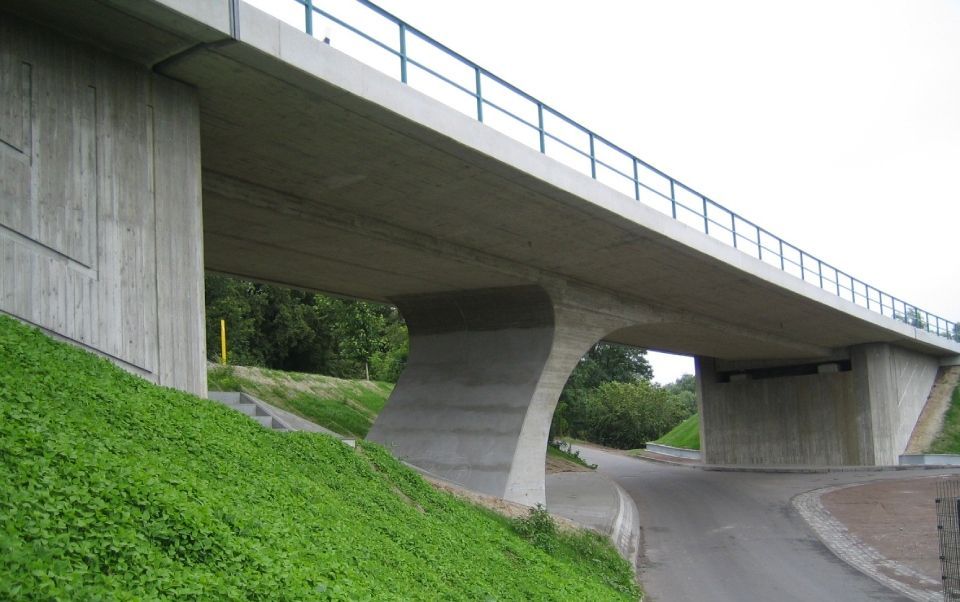 Betonüberführung einer Autobahn, die eine kurvige Straße neben einem leuchtend grünen, grasbewachsenen Damm überspannt