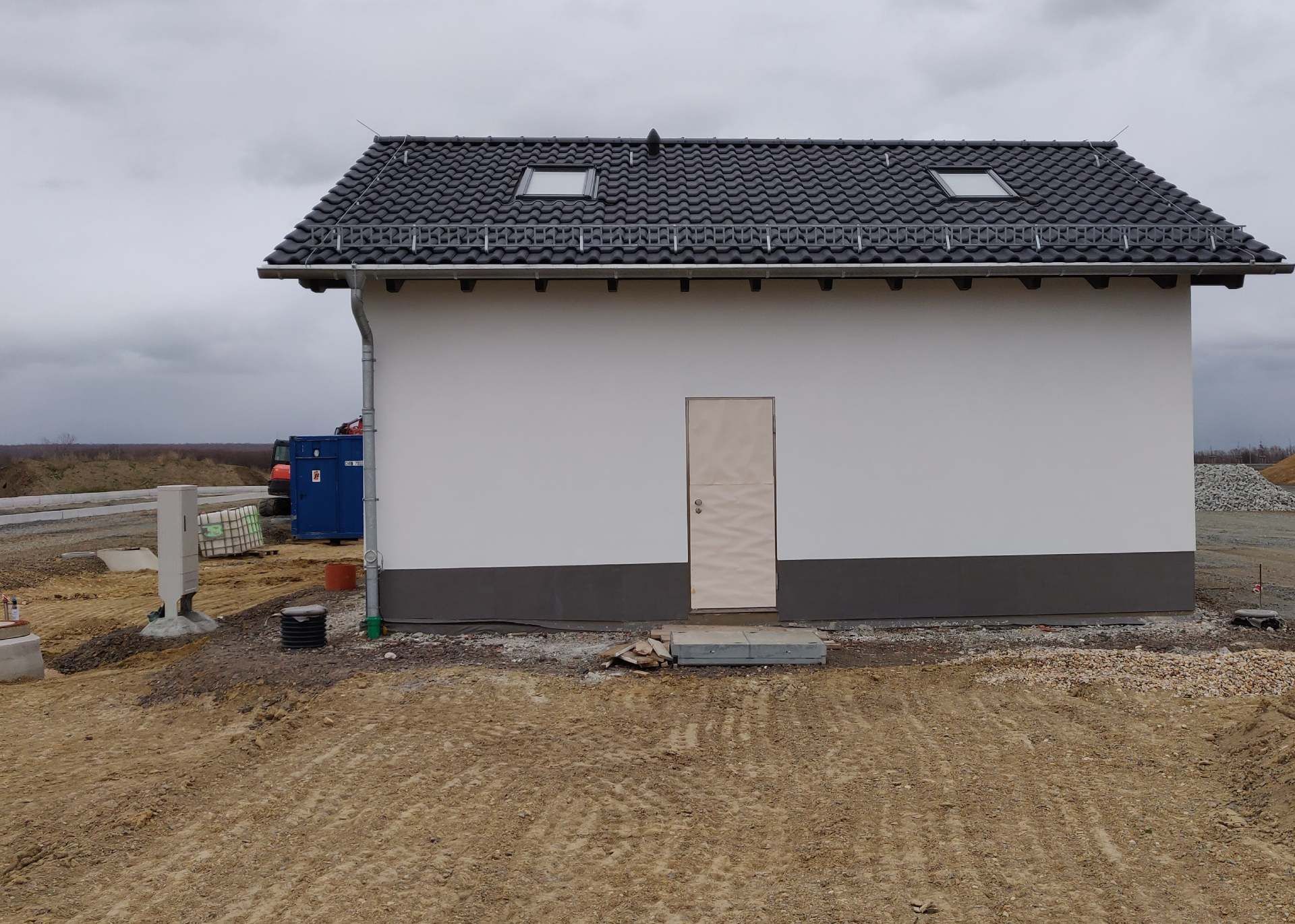 Kleines, unfertiges Haus mit grauem Dach und weißen Wänden auf einer schlammigen Baustelle.