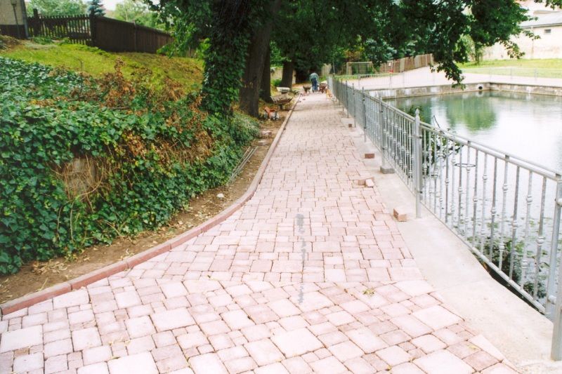 Gepflasterter Parkweg neben einem Teich, gesäumt von Grünpflanzen und einem Metallgeländer