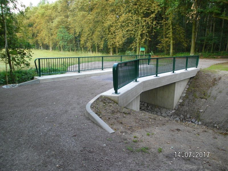 Kleine, mit grünem Geländer versehene Fußgängerbrücke über einen Schotterweg in einem bewaldeten Park