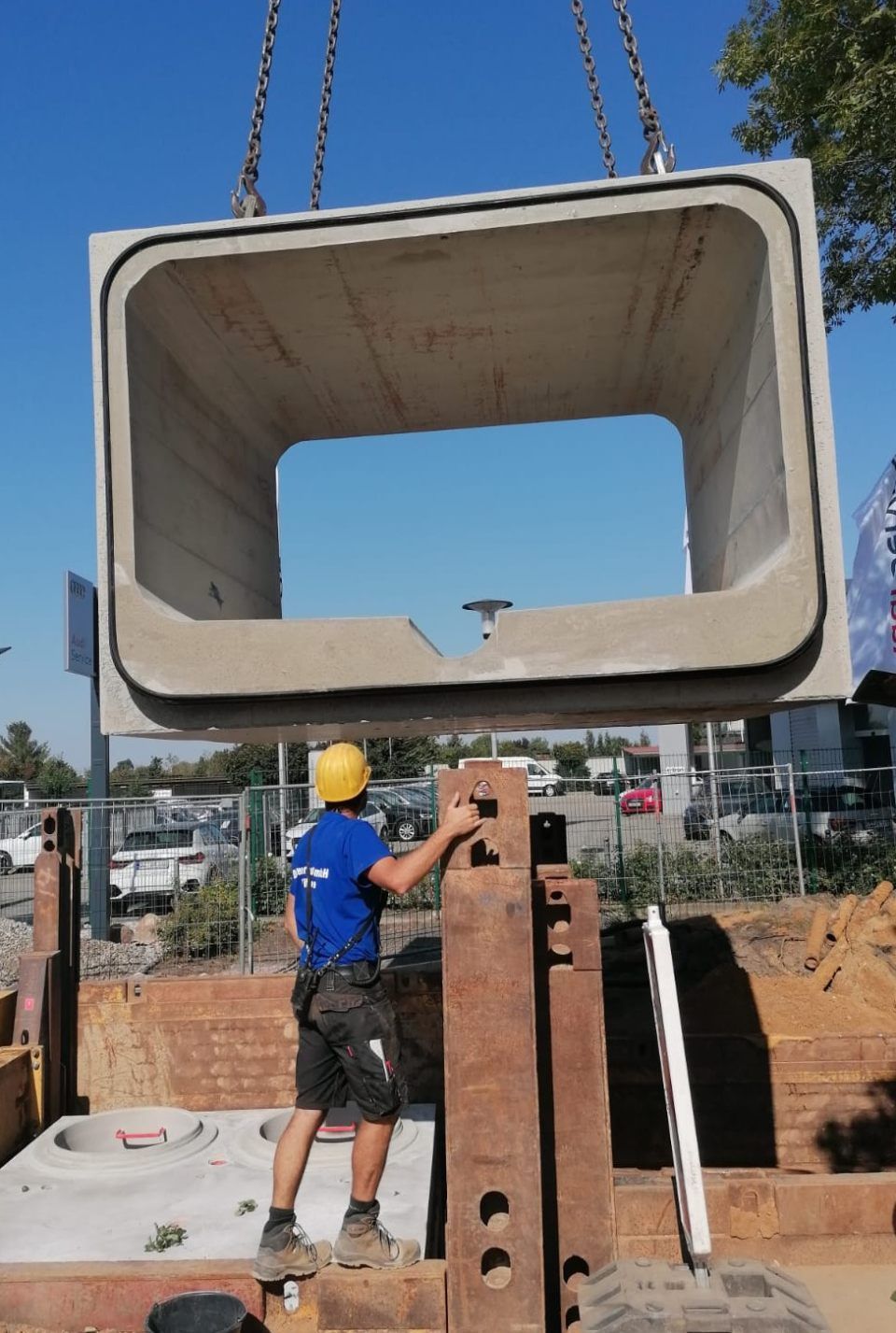 Ein Arbeiter mit gelbem Schutzhelm installiert mit einem Kran einen großen Betonrohrdurchlass.