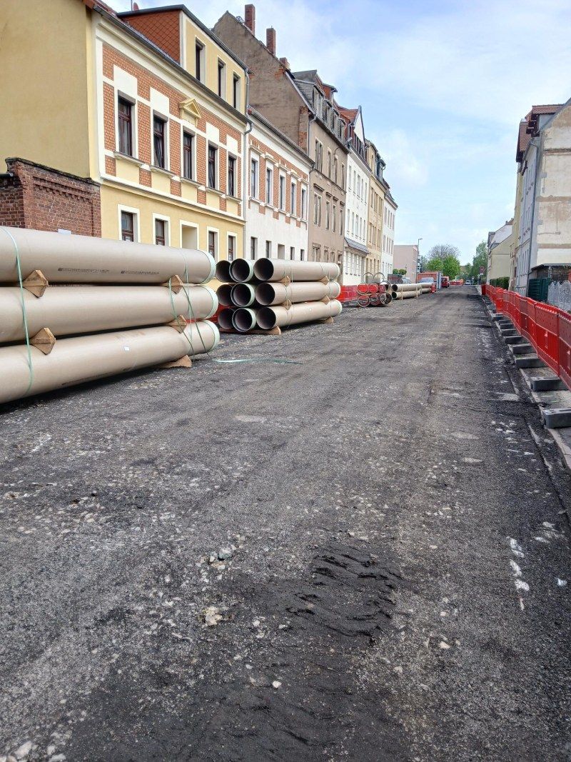 Eine schmale, im Bau befindliche Straße mit übereinander gestapelten Rohren, Schotterfahrbahn und Gebäuden auf beiden Seiten.