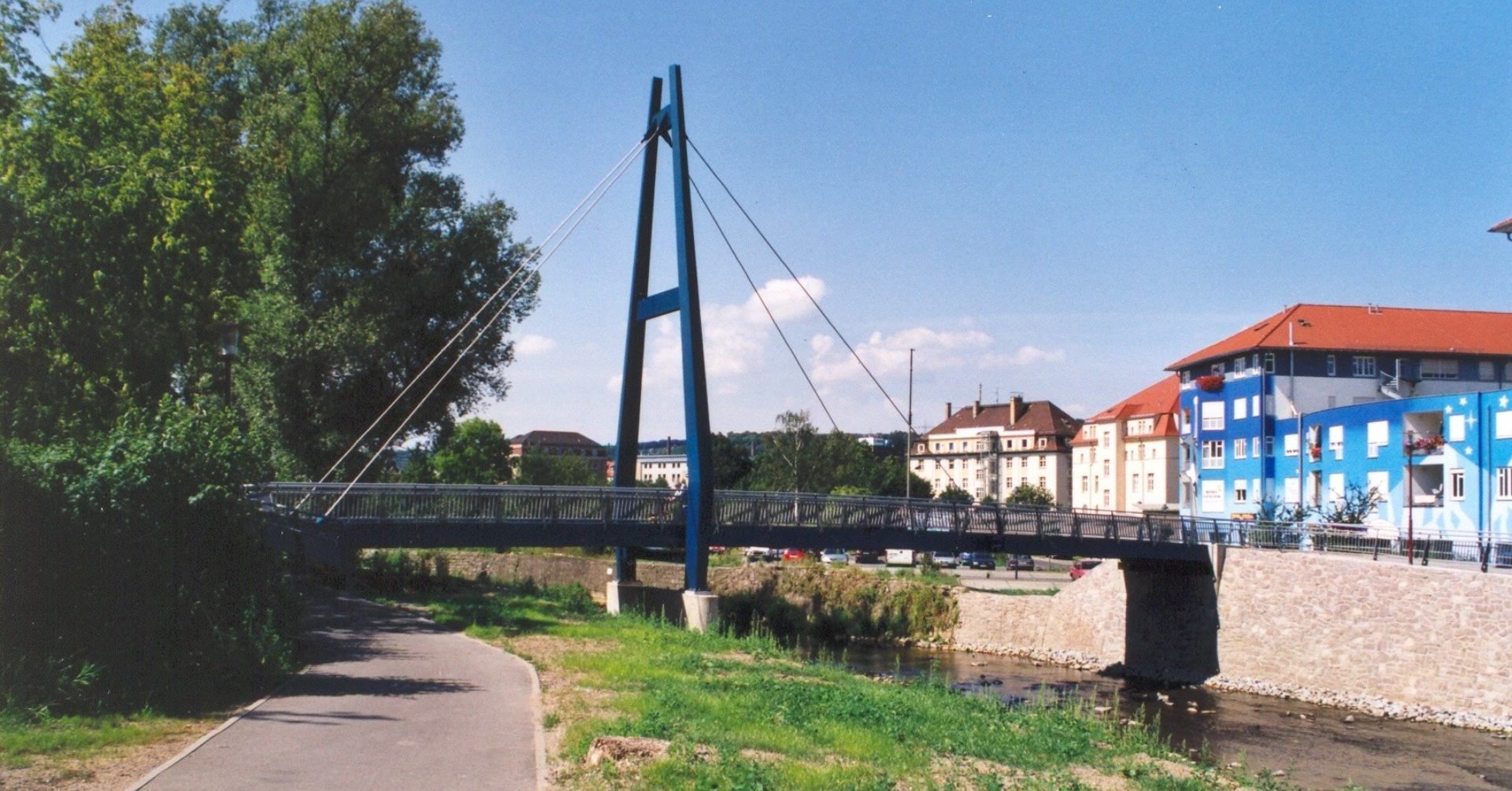 Moderne Hängebrücke über einen Fluss neben einem Weg, im Hintergrund blaue und rote Gebäude