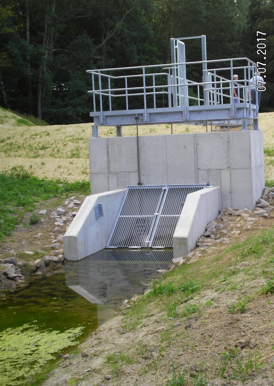 Betonbauwerk zur Wasserregulierung mit Metallgitter und Geländer neben einem grasbewachsenen Bach