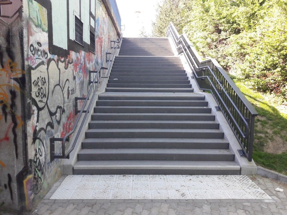 Außentreppe neben einer mit Graffiti bedeckten Mauer, rechts davon befinden sich Metallgeländer und Bäume.