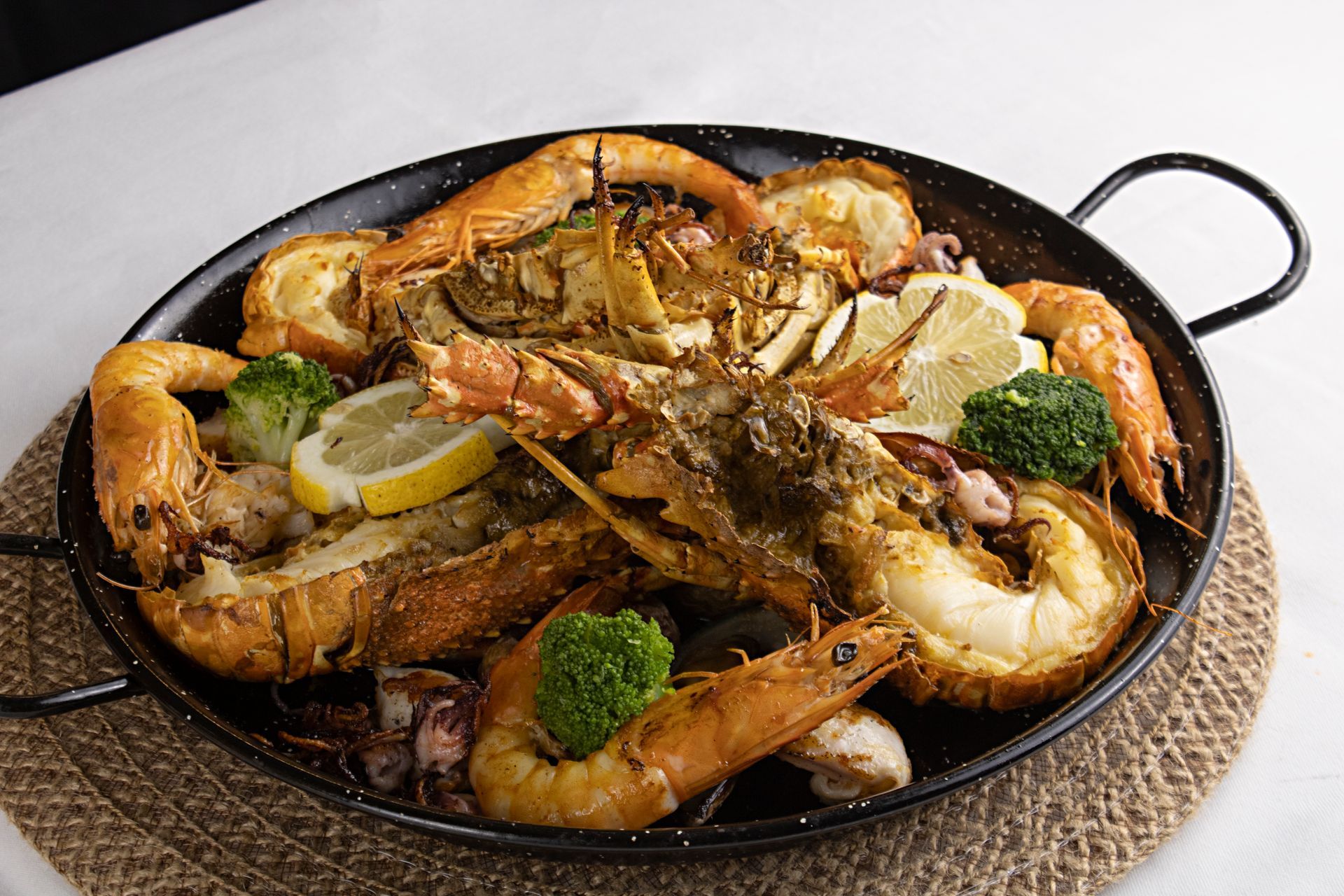 Paella de frutos do mar: lagosta, camarão, brócolis, fatias de limão em uma panela preta sobre uma esteira de tecido.