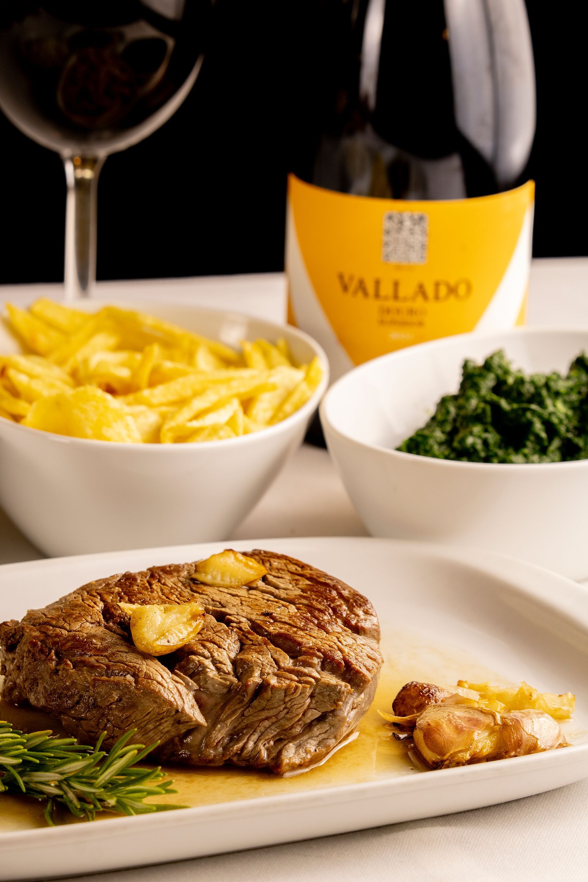 Bife com acompanhamentos de batatas fritas e espinafre, acompanhado de vinho tinto e uma garrafa de vinho Vallado.